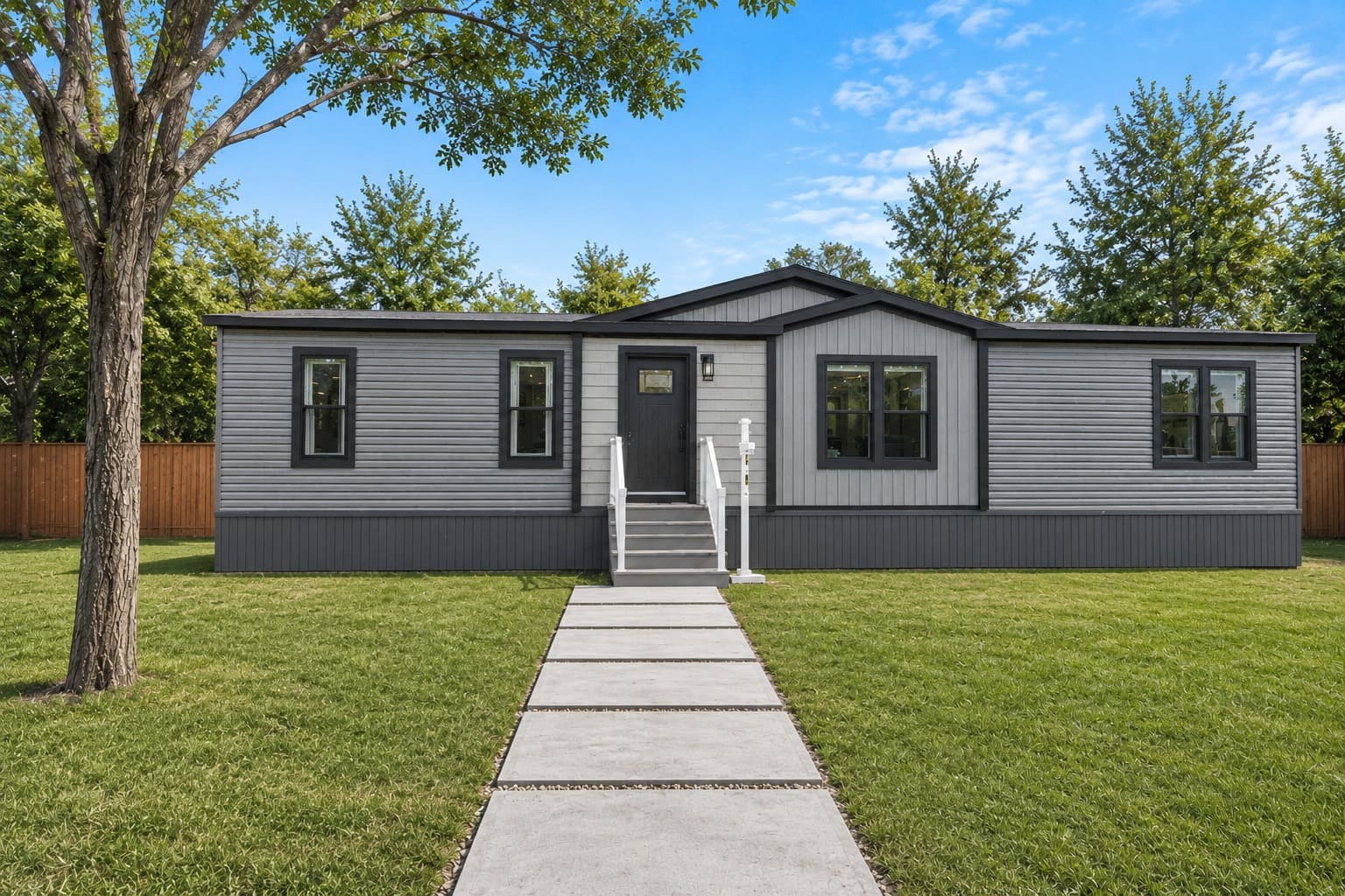 Gray board-and-batten exterior of a Lucky Ranch manufactured home — central gable, white front door with white-railed steps, paver-stripe walkway through manicured grass, dark trim windows
