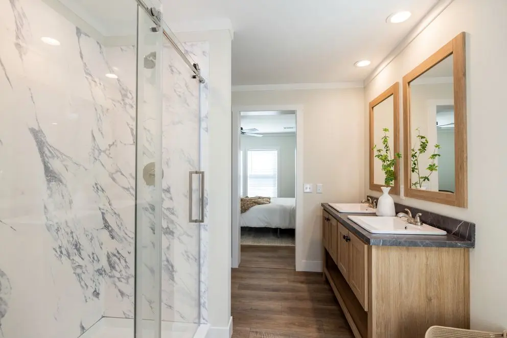 Ensuite bathroom — full-height marble walk-in shower with sliding glass door, dual vanity with stone countertop and natural wood cabinets, framed mirrors, and view through to the bedroom