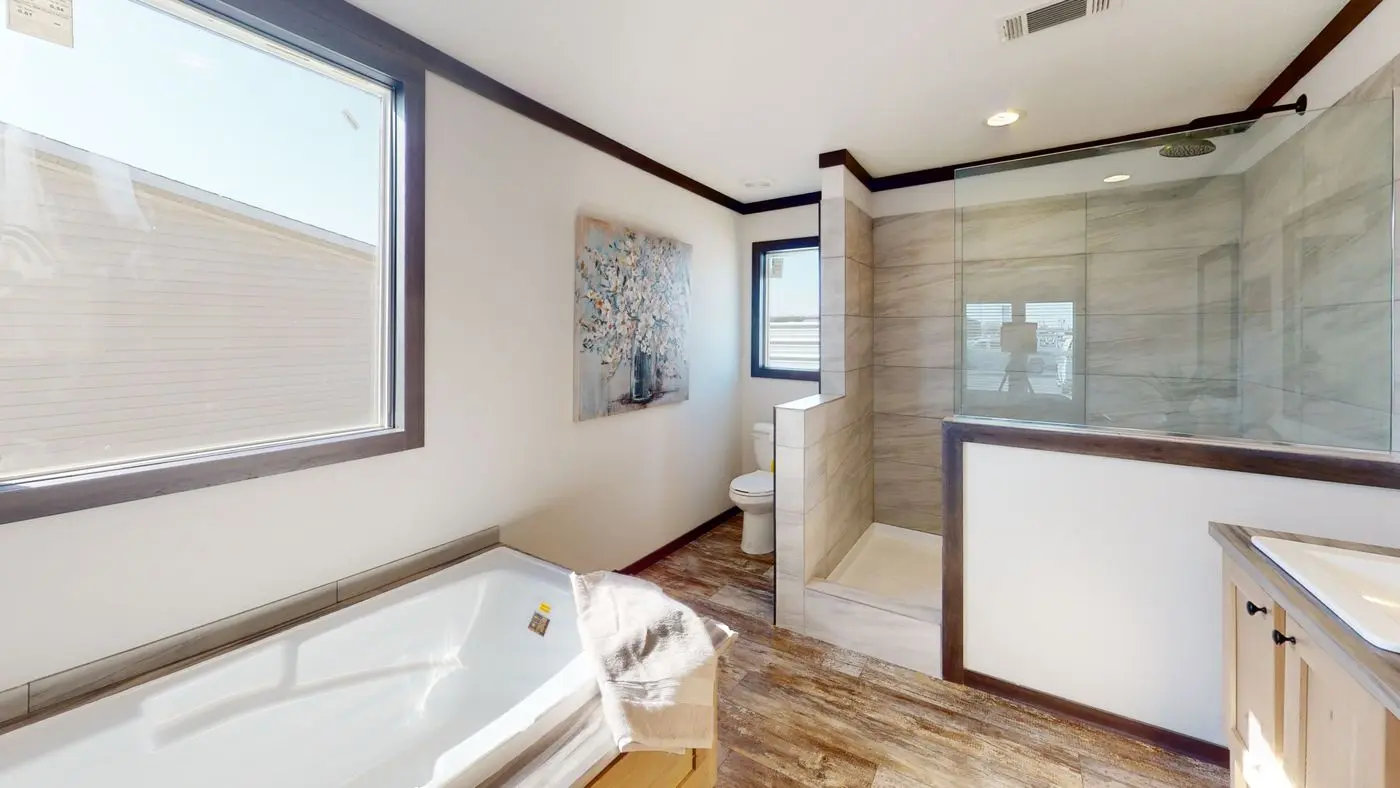 Master bathroom — deep soaking tub beneath a wide window, walk-in tile shower with glass partition and rain head, wood-look plank flooring, and framed botanical art