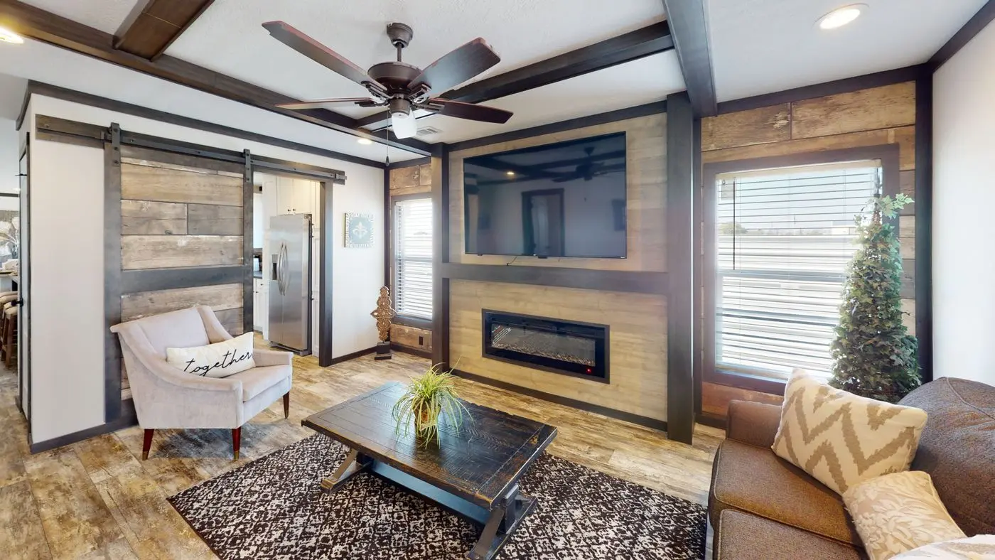 Living room of a Lucky Ranch model — coffered wood-beam ceiling, wood-clad fireplace wall with mounted TV and electric insert, sliding barn door pantry, brown sectional, accent chair, and patterned area rug