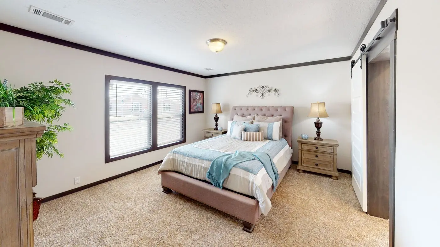 Master bedroom — cream walls, upholstered headboard, layered blue and neutral bedding, warm bedside lamps, plush carpet, and a sliding barn door to the ensuite